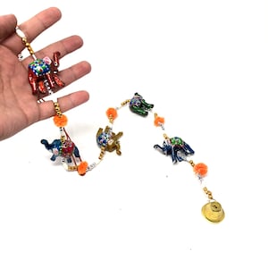 Könnte beinhalten: Eine Schnur mit bunten, handbemalten Elefantenfiguren mit goldenen Akzenten, die mit weißen Perlen und orangefarbenen Bommeln besetzt ist. Eine goldene Glocke hängt am Ende der Schnur.