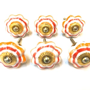 May include: Six white ceramic cabinet knobs with orange and red stripes and a gold center. The knobs have a scalloped edge.