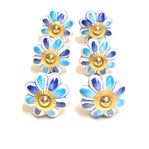 May include: Six ceramic flower-shaped cabinet knobs. Each knob has a white base with blue and light blue petals, and a gold-coloured centre. The knobs are arranged in two rows of three against a white background.