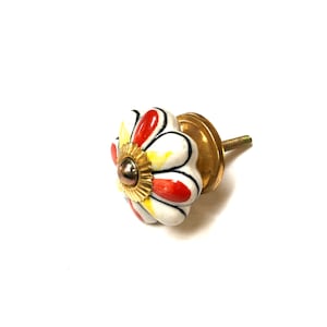 May include: White ceramic cabinet knob with a red and yellow flower design. The knob has a gold metal base and screw.