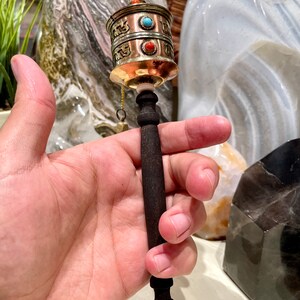 May include: A small handheld prayer wheel with a copper and brass body, adorned with turquoise and coral accents. The wheel is attached to a dark wooden handle.