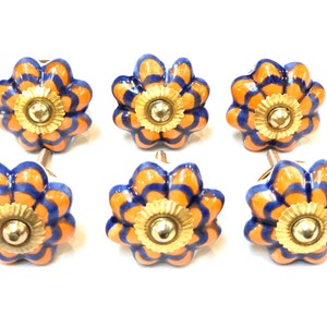 May include: Six ceramic drawer knobs in the shape of a flower with orange and blue stripes. The center of each knob has a gold-colored ring with a small, round, silver knob.