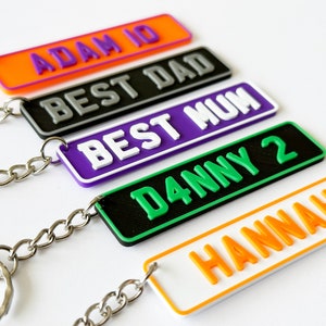 May include: A selection of keyrings with custom text in vibrant colours. Each keyring features a rectangular design with rounded edges and a metal chain. The text includes names and phrases such as "ADAM 10", "BEST DAD", "BEST MUM", "DANNY 2", and "HANNAH".