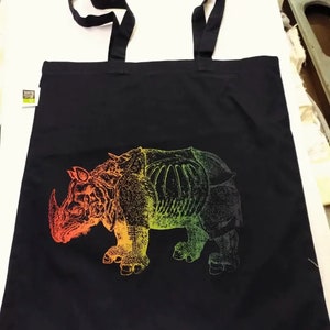 May include: Black tote bag with a colorful rhino graphic. The rhino design transitions from red to yellow to green. The bag has black handles and is made of a sturdy material, suitable for carrying groceries or other items.