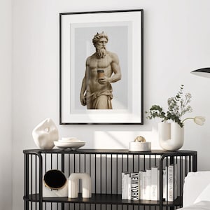 Könnte beinhalten: Gerahmter Kunstdruck mit einer klassischen Statue, die eine Kaffeetasse hält. Das Kunstwerk wird über einem schwarzen Konsolentisch mit dekorativen Objekten und Büchern ausgestellt. Der Raum ist in neutralen Tönen mit einer modernen Stehlampe eingerichtet.