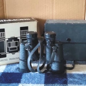 May include: A pair of black Simmons prism binoculars with a black carrying case. The binoculars have a textured finish and are in focus.