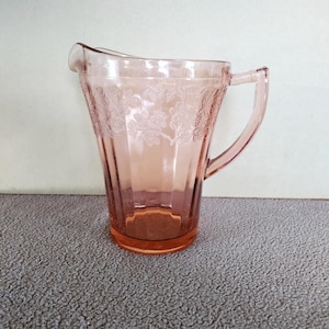 May include: A translucent pink glass pitcher featuring a molded floral pattern around the upper rim and a series of vertical fluted panels extending down the body. The vessel includes a curved handle and a small pouring spout at the top edge.