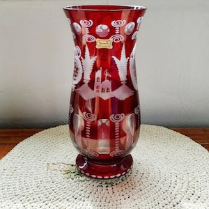 May include: A ruby red glass vase with intricate white etched designs. The vase features a flared top, a wide body, and a circular base. The etched patterns include swirling motifs, architectural elements, and a decorative label. A decorative piece for the home.