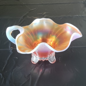 Puede incluir: Un cuenco de vidrio iridiscente con borde ondulado y asa. El cuenco tiene un brillo vibrante y multicolor, con tonos de naranja, amarillo y verde. Se asienta sobre una base decorativa de vidrio transparente con detalles ornamentados.