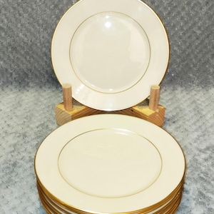 Op de afbeelding: Een set van zes ronde, crèmekleurige keramische borden met een dunne, metallic gouden rand. Eén bord staat rechtop op een houten standaard, terwijl de overige vijf borden eronder in een nette, gelijkmatige stapel zijn geplaatst.