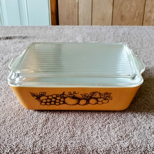 Puede incluir: Un plato para hornear Pyrex vintage con una tapa de vidrio transparente. El plato es amarillo con un diseño floral marrón de uvas y hojas.