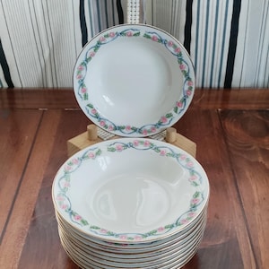 以下が含まれることがあります： 白いセラミックボウルのセット。各ボウルは、ピンクのバラと青いリボンのボーダーと金色の縁取りが特徴です。ボウルは木製の表面に積み重ねられています。