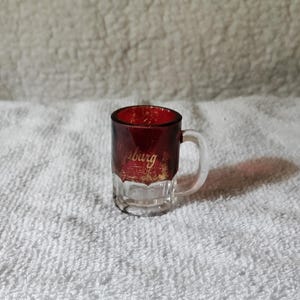 Op de afbeelding: Een klein, rood glazen mok met een gouden inscriptie die "Ypsilburg Is" luidt. De mok heeft een handvat en een heldere glazen bodem.