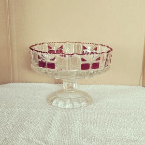 May include: A clear glass pedestal bowl with a scalloped rim and a red border. The bowl has a decorative pattern and a round base. The bowl is likely used for serving desserts or displaying decorative items.