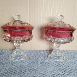 Può includere: Due bomboniere in vetro rosso rubino con coperchi. I piatti hanno una base a piedistallo e un coperchio in vetro trasparente con un pomello rotondo. Il vetro ha un motivo dettagliato attorno alla base e al coperchio. I piatti sono su una superficie blu.