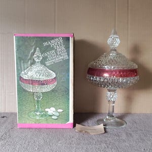 Può includere: Un piatto per caramelle in vetro trasparente con una fascia rossa attorno alla parte superiore. Il piatto è su un piedistallo e presenta un motivo a diamante. Il piatto è nella sua scatola originale, che è rosa e bianca e ha il testo "Diamond Point Tall Candy Box Ruby Band Crystal 0203 Indiana Glass Co Dunkirk Indiana 47236" stampato su di essa.