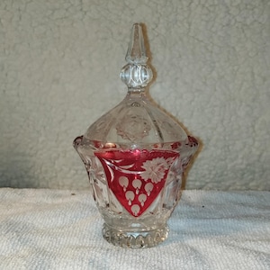 Puede incluir: Un azucarero de vidrio transparente con un diseño floral rojo y blanco. El tazón tiene una tapa con un pomo en la parte superior.