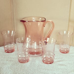 Puede incluir: Un juego de cuatro vasos de vidrio rosa y una jarra de vidrio rosa a juego. Los vasos tienen una base redondeada y una parte superior acampanada. La jarra tiene un asa y un pico.