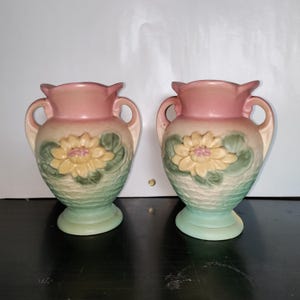 May include: A pair of ceramic vases featuring a gradient color scheme transitioning from a soft pink rim to a pale yellow and green base. Each vase is adorned with a raised, sculpted yellow water lily flower and green leaves on the front.