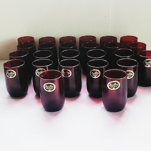 May include: A collection of twenty-four ruby-colored stemless glasses. Each glass has a rounded shape and a gold label. The glasses are arranged in neat rows on a white surface, creating a visually appealing display.
