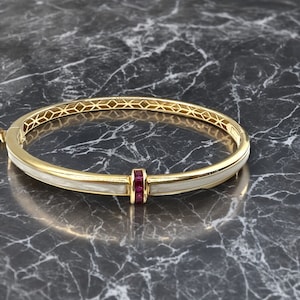 May include: A gold bangle bracelet with a white enamel band and a row of red ruby stones.