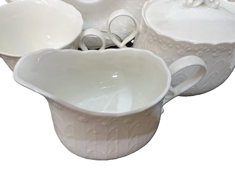 May include: A white ceramic tea set featuring a creamer, sugar bowl, and teacups. The pieces have a detailed embossed floral design and curved handles. The set is suitable for serving tea or coffee.