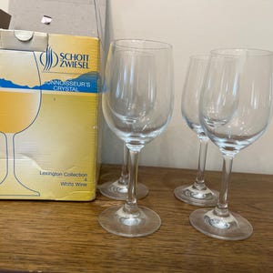K&ouml;nnte beinhalten: Vier klare Weingl&auml;ser aus Glas, von denen zwei mit Wei&szlig;wein gef&uuml;llt sind. Die Gl&auml;ser stehen auf einer Holzoberfl&auml;che. Die Schachtel im Hintergrund ist mit "Schott Zwiesel Connoisseur's Crystal Lexington Collection 4 White Wine" beschriftet.
