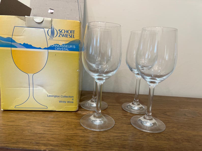 K&ouml;nnte beinhalten: Vier klare Weingl&auml;ser aus Glas, von denen zwei mit Wei&szlig;wein gef&uuml;llt sind. Die Gl&auml;ser stehen auf einer Holzoberfl&auml;che. Die Schachtel im Hintergrund ist mit "Schott Zwiesel Connoisseur's Crystal Lexington Collection 4 White Wine" beschriftet.