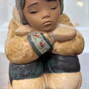 May include: A ceramic figurine of a young person wearing a brown and blue winter coat and hat, sitting with their arms crossed and a sad expression.