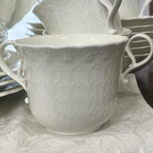 May include: A white teacup with two handles, part of a matching tea set. The cup has a detailed embossed pattern and sits on a white patterned surface. Other teacups, saucers, and plates are visible in the background.