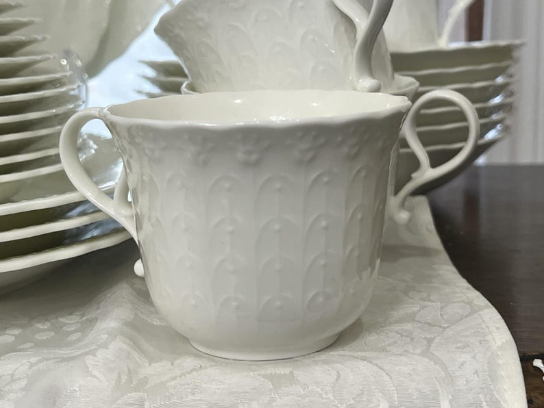 May include: A white teacup with two handles, part of a matching tea set. The cup has a detailed embossed pattern and sits on a white patterned surface. Other teacups, saucers, and plates are visible in the background.