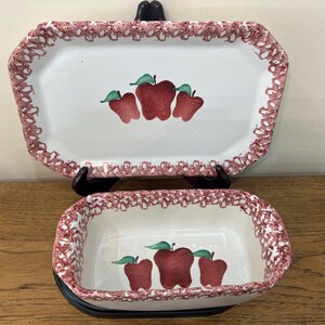 Puede incluir: Dos platos de cerámica con un diseño de manzanas rojas y blancas. El plato más grande es rectangular y el más pequeño es ovalado. Ambos platos tienen un borde festoneado.