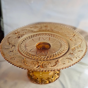 May include: Amber glass cake stand with a decorative, scalloped edge and a raised, floral design. The stand has a round base with a scalloped edge.