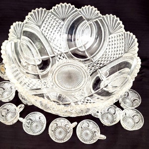 May include: A vintage crystal punch bowl and 10 cups with intricate cut glass designs. The bowl has a scalloped edge and a large central design. The cups have a similar design with a small handle.
