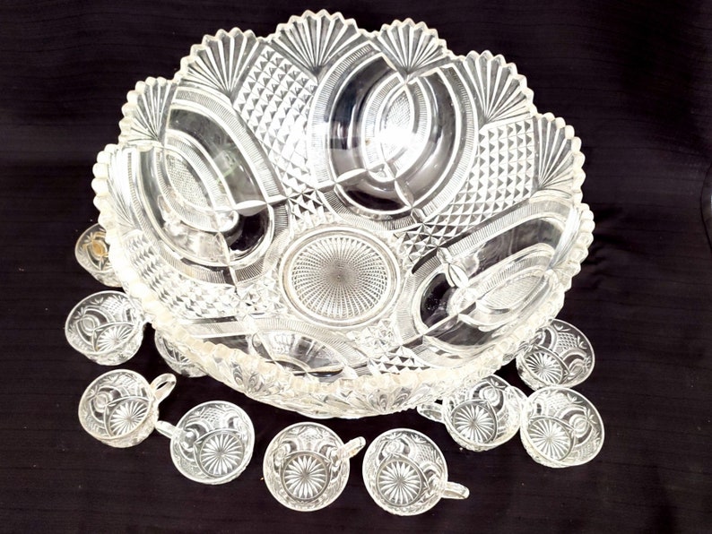 May include: A vintage crystal punch bowl and 10 cups with intricate cut glass designs. The bowl has a scalloped edge and a large central design. The cups have a similar design with a small handle.