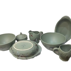 Puede incluir: Una colección de fuentes de cerámica verde salvia. El juego incluye cuencos, una fuente cubierta, una fuente para servir y jarras. Cada pieza presenta un delicado diseño de pájaros y flores. Los platos están dispuestos sobre un fondo blanco.