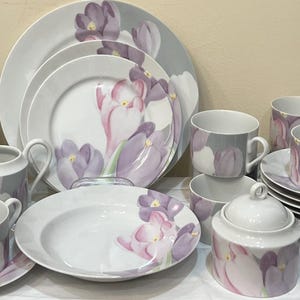 May include: A complete white porcelain dinnerware set featuring a floral design with purple and white crocus flowers. The set includes plates, bowls, cups, saucers, a creamer, and a sugar bowl. The plates are stacked in multiple piles.