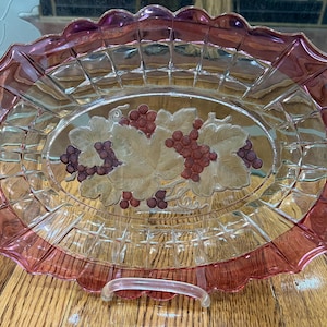 Op de afbeelding: Een ovale serveerschaal van helder glas met een roze rand en een druivenrankenontwerp. De schaal heeft een reliëf, getextureerd patroon.