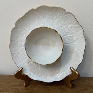 Può includere: Piatto da portata in ceramica bianca con bordo dorato e una ciotola bianca più piccola con bordo dorato all'interno. Il piatto ha un design a foglie in rilievo.