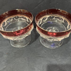 Puede incluir: Dos cuencos de postre de cristal transparente con un borde ancho de color rojo intenso y una base festoneada texturizada. Cada cuenco tiene un tallo corto y robusto y una base circular, con un patrón de puntos en relieve en la parte superior.