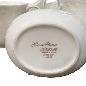 May include: White bone china serving pieces, including a creamer and a cup, with embossed floral patterns. The underside of the creamer displays the text "Bone China Mikasa White Silk A7050 Made in Japan."