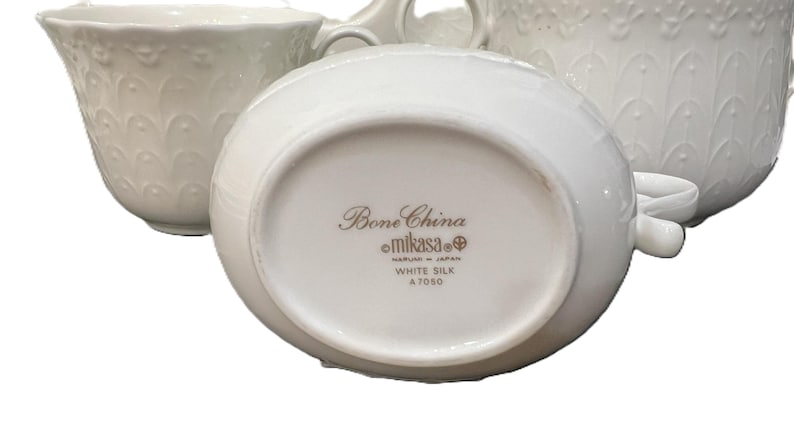 May include: White bone china serving pieces, including a creamer and a cup, with embossed floral patterns. The underside of the creamer displays the text "Bone China Mikasa White Silk A7050 Made in Japan."