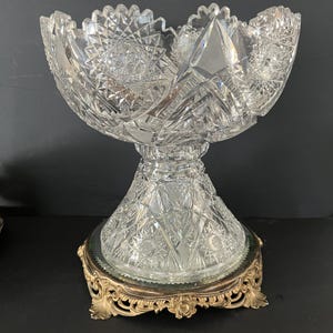 May include: A clear, cut-glass bowl with a pedestal base, set on an ornate gold-toned metal stand. The bowl features intricate geometric patterns and a scalloped rim, showcasing a classic design. The stand adds an elegant touch to the overall presentation.