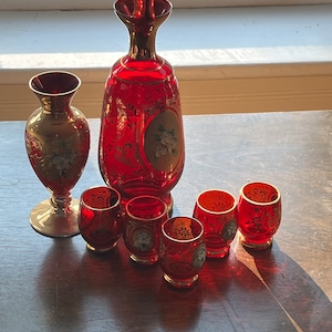Puede incluir: Un juego de jarras y vasos de chupito de vidrio rojo con detalles dorados y diseños florales. La jarra tiene un tapón dorado.