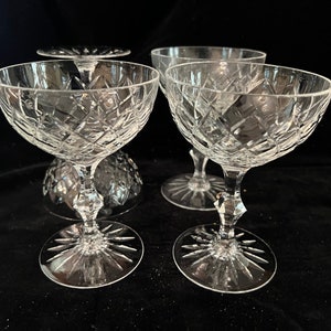 May include: Four clear glass champagne coupes with a cut glass design. The coupes are arranged on a black surface.