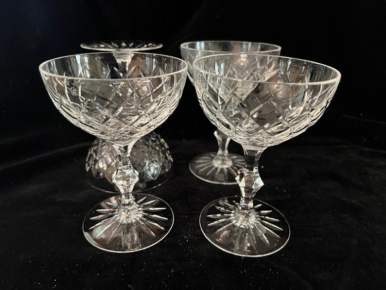 May include: Four clear glass champagne coupes with a cut glass design. The coupes are arranged on a black surface.