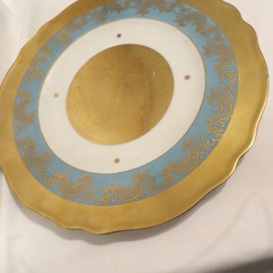 Könnte beinhalten: Ein goldener und blauer Dekorationsteller mit gewelltem Rand. Der Teller hat eine weiße Mitte mit einem goldenen Rand und einem blauen Band mit einem goldenen Blumenmuster.