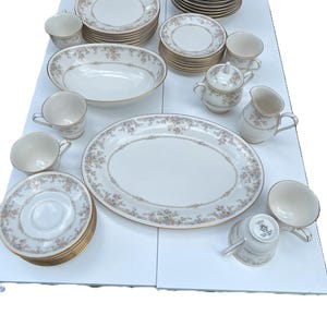 Puede incluir: Un juego completo de vajilla de porcelana vintage. El juego incluye platos, cuencos, tazas, platillos, una fuente para servir, una azucarera, una jarra de crema y una jarrita. Las piezas son blancas con un estampado floral y ribete dorado.