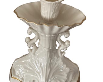 May include: A white ceramic candlestick holder with a scalloped dish and a floral-shaped candle cup. The base features a leaf design and gold trim. The candlestick holder is approximately 7 inches tall.