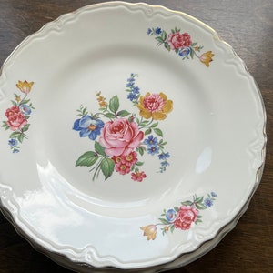May include: A round, off-white ceramic dinner plate with a scalloped edge and a thin gold rim. The plate features a central floral bouquet of pink, yellow, and blue flowers with green leaves, complemented by two smaller matching floral sprays on the rim.